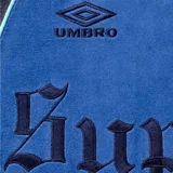 Campera deportiva de terciopelo Supreme x Umbro en color azul, con cuello alto, cierre frontal completo y bolsillos laterales. Cuenta con paneles en distintos tonos de azul y celeste, líneas curvas decorativas, y los logotipos de Umbro y Supreme bordados en el pecho junto con el escudo del Club Nacional de Football.