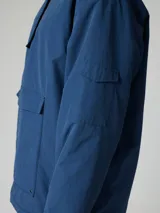 Canguro tipo pullover de color azul, con capucha ajustable, bolsillo frontal tipo canguro y cierre parcial en el cuello. Presenta un diseño funcional con puños elásticos y un bolsillo adicional en la manga.