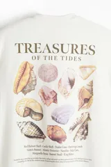 Remera de algodón blanca de corte holgado, con cuello redondo y diseño estampado en el pecho con las palabras 'Treasures of the Tides' y una pequeña ilustración de un caracol marino.