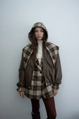 Chaqueta de cuero sintético color marrón con capucha y solapas amplias en tela escocesa a cuadros. Posee cierre frontal, puños elásticos y diseño de corte holgado.