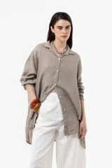 Camisa de lino color beige, de calce amplio y relajado, con cuello camisero, cierre frontal de botones, mangas largas y diseño de largo irregular con aberturas laterales.