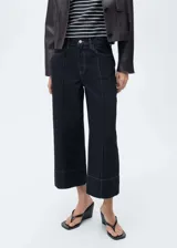 Pantalón de jean estilo wide leg con tiro medio y diseño crop. Presenta costuras verticales decorativas en el frente, cinco bolsillos, trabillas para cinturón y cierre frontal con cremallera y botón.