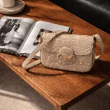 Cartera tipo bandolera de cuero con textura grabada, color beige. Presenta un detalle de herraje metálico dorado en la solapa frontal y placa con logo de la marca. Incluye correa ajustable para llevar al hombro o cruzada.