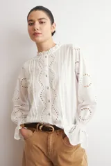 Camisa blanca de manga larga con cuello alto y volados, confeccionada en tela con bordado inglés y detalles calados verticales. Presenta puños con volados y cierre frontal con botones.