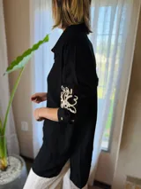 Camisa de manga larga confeccionada en muselina negra, con diseño de bordado artesanal en color crema sobre la manga. Presenta cuello clásico y cierre frontal con botones.
