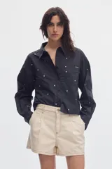 Camisa cropped de manga larga negra con estampado de lunares blancos, cuello camisero, cierre frontal de botones y bolsillos plaqué en el pecho.