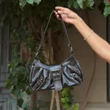 Cartera bandolera negra de cuero sintético con correa de hombro ajustable. Presenta un cierre con hebilla plateada en la solapa frontal y detalles de hebillas decorativas en los laterales.