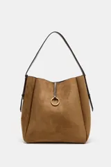 Bolso tipo saca de gran tamaño en color marrón, confeccionado en material efecto gamuza. Presenta un diseño minimalista con una anilla metálica decorativa en el frente, asa de hombro ajustable y cierre mediante botón imantado. Incluye bolsillo interior con cremallera.