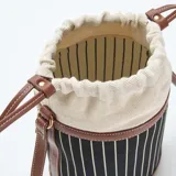 Bolso tipo saco con cuerpo de tela a rayas verticales en blanco y negro, con detalles de cuero sintético color marrón en la base, borde superior y correa ajustable.
