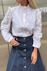 Camisa de algodón con estampado de rayas finas, cuello alto con detalle de broderie y mangas 3/4 con frunces en los hombros y apliques de encaje. Presenta un diseño con jaretas frontales y cierre de botones.