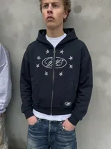 Campera de abrigo estilo canguro negra con cierre frontal completo, capucha y bolsillos tipo canguro. Presenta un estampado frontal con tipografía en un óvalo rodeado de estrellas blancas y un pequeño logo en la parte inferior.