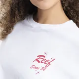 Remera blanca de corte oversized, con cuello redondo y mangas cortas. Presenta un pequeño estampado en el pecho con el logo de la marca Reef en color rojo y detalles de estrellas.