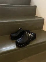 Zuecos negros de cuero encerado con detalle de tachas plateadas en la base y hebilla metálica en el empeine.