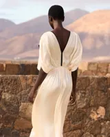 Vestido largo color crudo, confeccionado en viscosa satinada de inspiración grecorromana. Cuenta con escote en V profundo con ribete negro en contraste, mangas cortas fluidas con detalles oscuros, y corte en la cintura con drapeado suave.