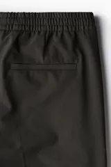 Pantalón tipo jogger de vestir color marrón oscuro, con cintura elástica y cordón ajustable. Presenta pliegues marcados en la parte delantera y un corte slim fit.
