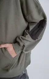 Hoodie oversize color verde militar, confeccionado en algodón rústico. Presenta capucha forrada, bolsillo frontal tipo canguro y un bolsillo adicional con cierre en la manga izquierda, con detalles en microfibra negra.