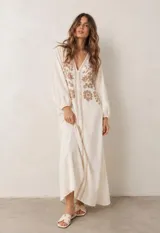 Vestido largo beige con escote en V y mangas largas abullonadas, adornado con delicados bordados florales en tono tostado en la parte frontal y cenefas decorativas a lo largo de la abertura central.