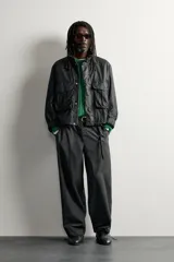 Cazadora regular fit de Zara en colaboración con Aaron Levine, confeccionada en tejido de algodón con acabado efecto encerado en color negro. Presenta cuello subido, manga larga con puño ajustable, bolsillos frontales con solapa y cierre de cremallera oculto con solapa de botones a presión.