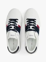 Zapatillas urbanas Tommy Hilfiger de corte bajo, confeccionadas en piel lisa color blanco. Presentan paneles laterales en azul marino y rojo, cordones oscuros y suela de goma gruesa con diseño dentado. Incluyen detalles de la marca en la lengüeta, lateral y talón.
