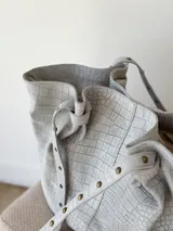 Cartera tipo shopper de cuero gris con textura símil cocodrilo, diseño amplio, correas ajustables con tachas y cierre superior.