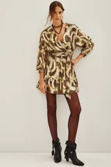 Vestido corto de estilo wrap con estampado abstracto en tonos dorados y marrones con acabado metalizado. Presenta escote en V cruzado, mangas largas con puños elásticos, lazo ajustable en la cintura y falda con volado en el ruedo.
