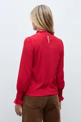Blusa de color rojo con cuello alto y detalle de volados, mangas largas con puños elásticos en punto smock y textura sutil en la tela.