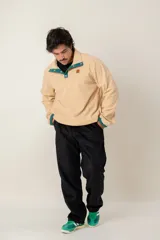 Buzo de pana color beige, de corte oversize, con cuello alto y cierre frontal parcial con botones a presión. Presenta detalles en color verde en la tapeta de los botones y en los puños, además de un parche decorativo en el pecho.