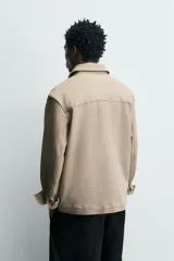 Sobrecamisa relaxed fit color beige con tejido elástico, cuello solapa, manga larga con puños abotonados, bolsillos de plastrón con solapa en el pecho, bolsillos laterales de vivo en la cadera y cierre frontal de botones.