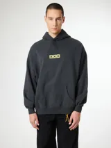 Hoodie gris oscuro con capucha, corte boxy y bolsillo tipo canguro. Presenta un pequeño estampado frontal con tres cuadros amarillos que contienen cruces negras.