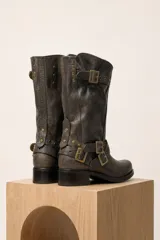 Bota de caña media de cuero color marrón oscuro, con diseño slouchy, hebillas metálicas decorativas y tachas alrededor del tobillo y en la parte superior. Cuenta con taco bajo y cuadrado.