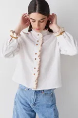Camisa de manga larga en color blanco, confeccionada en tejido de lino, con detalles de volados en crochet en el cuello, la botonadura frontal y los puños. Presenta detalles de costuras en color ocre en los hombros y puños, con cierre frontal de botones.