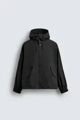 Cazadora técnica de corte relajado en color negro. Presenta cuello subido con capucha ajustable, cierre frontal con botones, mangas largas con puños elásticos y bolsillos frontales con solapa.