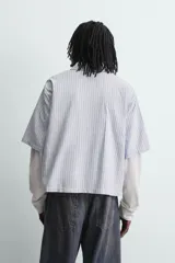Camisa de corte relajado con diseño de rayas verticales en tonos azul y blanco. Presenta cuello solapa, cierre frontal de botones y un detalle de texto bordado en color rojo en el pecho. Cuenta con un diseño de manga larga con efecto doble, combinando la manga corta de la camisa con una manga larga de tejido tipo waffle en color crudo.