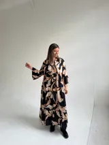 Vestido largo negro con estampado de hojas en tonos beige y tostado, de corte amplio y fluido, con escote en V y botonadura frontal parcial. Cuenta con mangas largas y volados en el faldón.