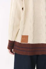 Buzo de tejido de punto con diseño de trenzas, color crema con detalles en marrón en cuello, puños y ruedo. Presenta líneas finas en contraste y pequeñas costuras decorativas en forma de cruz.