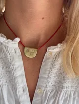 Collar con dije de bronce en forma de media luna con grabados de un trébol, una cruz y un sol. El collar es de hilo encerado rojo y tiene largo regulable.