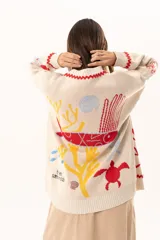 Saco tipo cardigan oversize de punto color beige, con diseño temático marino que incluye bordados de corales, peces, estrellas de mar y tortugas. Presenta detalles de galón picot en color rojo en los bordes y puños, cierre frontal con botones al tono y corte holgado.