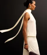 Blusa color crema sin mangas de crepé con cuello alto y lazo por tiras largas de tejido. La prenda tiene cierre por gota con detalle de perla artificial atrás del cuello y frunzidos en la parte delantera.