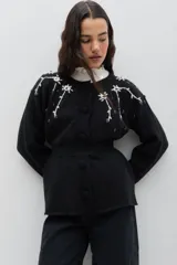 Saco de punto color negro con bordado floral artesanal en color blanco en la zona de los hombros. Presenta cuello redondo, cierre frontal con botones forrados en crochet y una banda elástica en la cintura que define la silueta.
