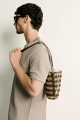 Bandolera vertical con acabado trenzado en tonos tostados y oscuros, cuenta con bolsa interior de tela con cierre de cordón ajustable y correa fina para el hombro.