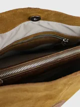 Cartera bandolera de cuero gamuzado color tostado, de diseño cuadrado, con correa larga ajustable y detalle de aplique circular con logo grabado en el centro.