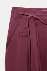 Pantalón de tiro alto color bordó, de corte ancho y pierna recta, con pinzas frontales y lazada ajustable en la cintura. El tejido presenta un acabado satinado o ligeramente arrugado.