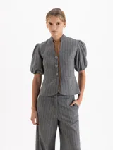 Chaqueta gris con diseño de rayas verticales y sutiles lentejuelas. Presenta cuello mao con lazo de gamuza, botones decorativos en forma de flor, recorte en la cintura y mangas abullonadas con puño elástico.