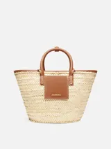 Bolso tipo canasto tejido a mano en fibra vegetal con detalles de cuero marrón, asas cortas enrolladas y logotipo Jacquemus en letras doradas en el frente.
