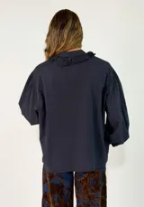 Camisa negra de corte holgado con cuello tipo bebé con volados y lazo de ajuste, cierre frontal con botones y mangas largas abullonadas con puños abotonados.