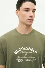 Remera verde militar de manga corta con cuello redondo y estampado frontal con el texto "Brookfield The Emporium of Postmodern Activity No 1001 Torino | 1971".