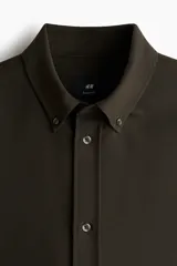 Camisa de manga larga color marrón oscuro, confeccionada en sarga suave, con cuello americano y tapeta clásica de botones. Presenta canesú en la espalda y bajo redondeado.