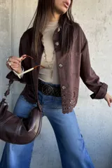 Chaqueta de gamuza color chocolate, con corte recto, cuello clásico y cierre frontal con botones. Presenta un diseño calado decorativo en el ruedo y bolsillos frontales tipo parche.