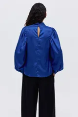 Blusa de tejido jacquard con acabado satinado y diseño floral en relieve. Presenta cuello elevado, mangas largas abullonadas con puños abotonados y cierre posterior con botones forrados.