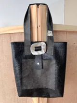 Bolso tote negro con textura símil cuero de cocodrilo, bolsillo frontal de cuero liso y cierre con botón metálico.